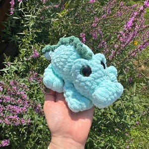 May include: A light blue crochet plush toy shaped like a small alligator or dinosaur. The creature features a textured, soft yarn body, a darker green ridged spine, and large, round, glossy black eyes positioned on the sides of its head.