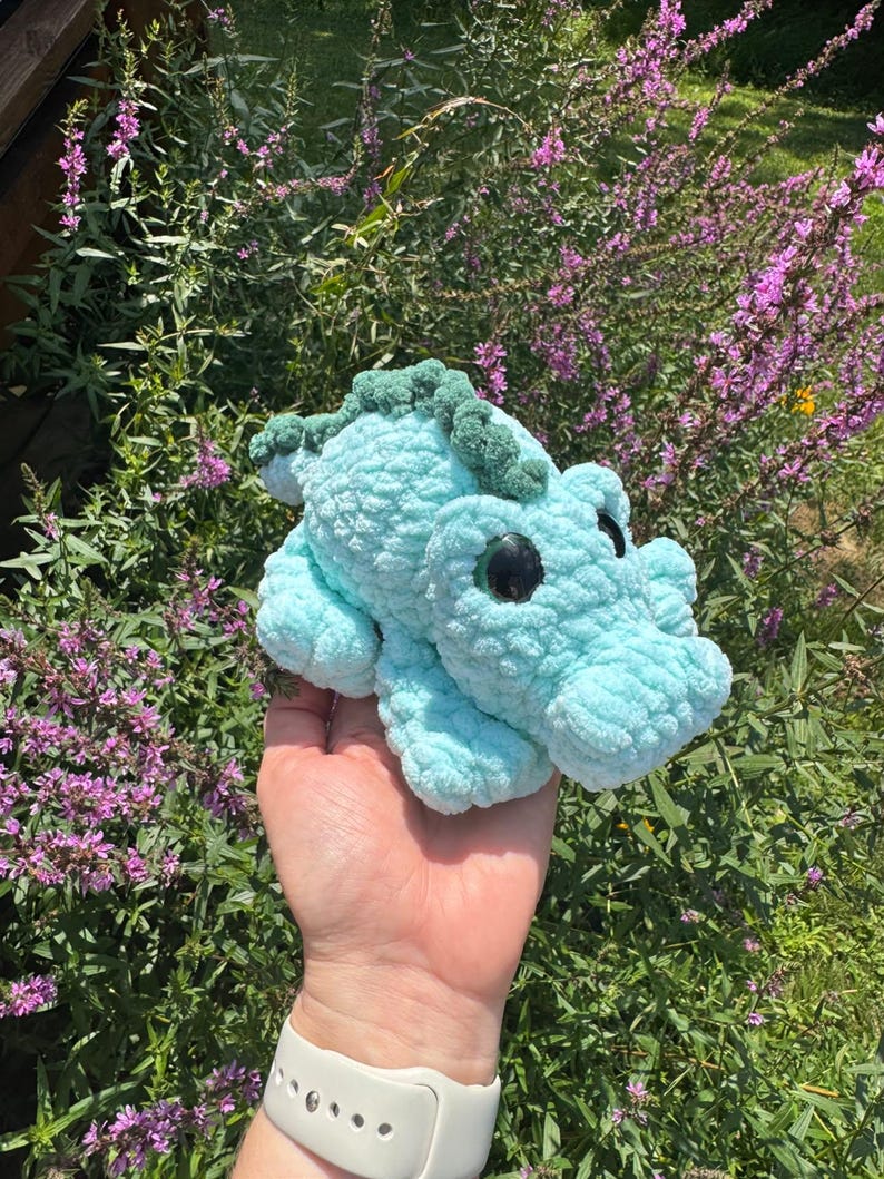 May include: A light blue crochet plush toy shaped like a small alligator or dinosaur. The creature features a textured, soft yarn body, a darker green ridged spine, and large, round, glossy black eyes positioned on the sides of its head.