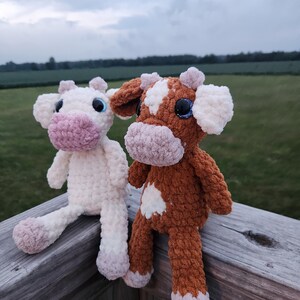 May include: Two crocheted stuffed cows, one white and one brown with white spots, sitting on a wooden railing. The cows have pink noses and blue eyes.