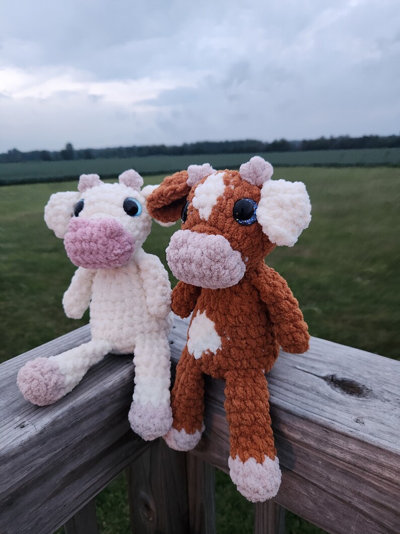 May include: Two crocheted stuffed cows, one white and one brown with white spots, sitting on a wooden railing. The cows have pink noses and blue eyes.