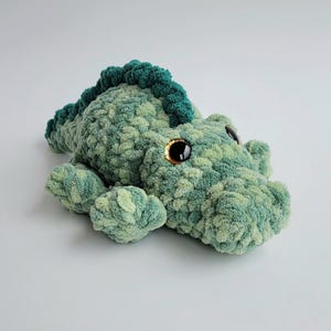 May include: A plush crocodile toy crafted from variegated green yarn with a textured, soft appearance. The stuffed animal features a dark green dorsal ridge, four short legs, and large, glossy black safety eyes with amber-colored irises.
