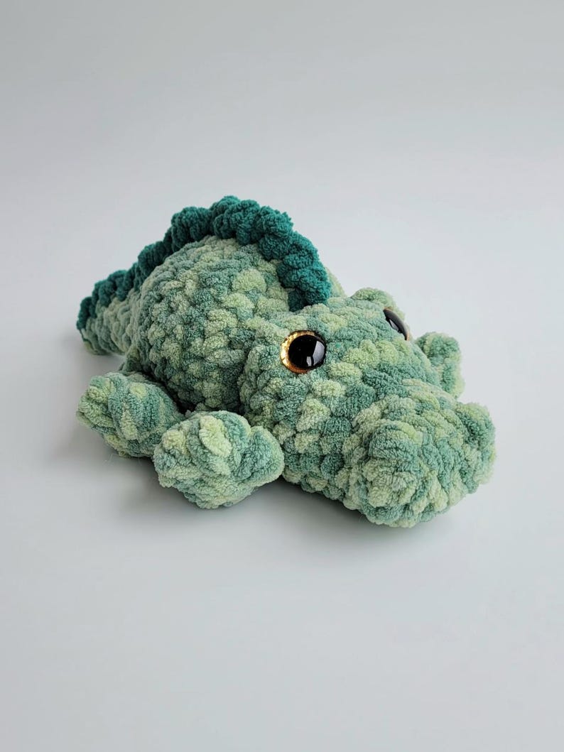 May include: A plush crocodile toy crafted from variegated green yarn with a textured, soft appearance. The stuffed animal features a dark green dorsal ridge, four short legs, and large, glossy black safety eyes with amber-colored irises.