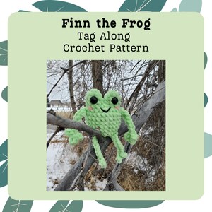 May include: A green crocheted frog with black eyes and a smiling mouth. The frog is sitting on a branch in a snowy forest. The image is titled "Finn the Frog Tag Along Crochet Pattern".