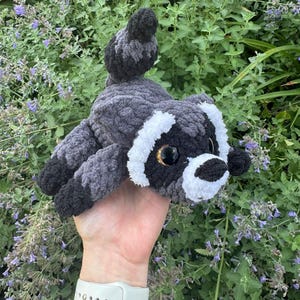 Op de afbeelding: Een knuffel in de vorm van een wasbeer, gemaakt van donkergrijs, lichtgrijs en wit garen. Het gehaakte speelgoed heeft een gestreepte staart, een zwart masker rond grote, glanzende, donkerbruine ogen en een witte snuit met een zwarte neus.
