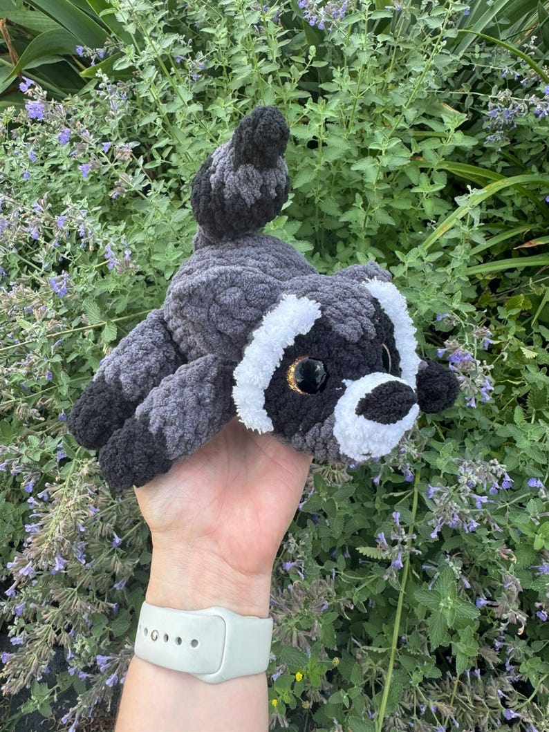 Op de afbeelding: Een knuffel in de vorm van een wasbeer, gemaakt van donkergrijs, lichtgrijs en wit garen. Het gehaakte speelgoed heeft een gestreepte staart, een zwart masker rond grote, glanzende, donkerbruine ogen en een witte snuit met een zwarte neus.