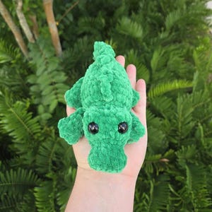 May include: A small, bright green plush alligator toy crafted from soft, textured yarn. The creature features a rounded snout, a ridged back, and two large, glossy black plastic eyes positioned on the top of its head.