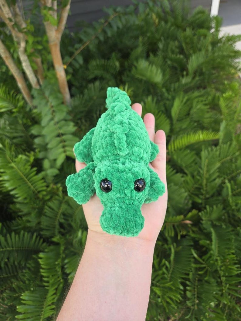 May include: A small, bright green plush alligator toy crafted from soft, textured yarn. The creature features a rounded snout, a ridged back, and two large, glossy black plastic eyes positioned on the top of its head.