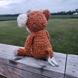 May include: A brown crocheted stuffed animal with white ears and a white tail sitting on a wooden railing. The animal is looking out at a field in the distance.