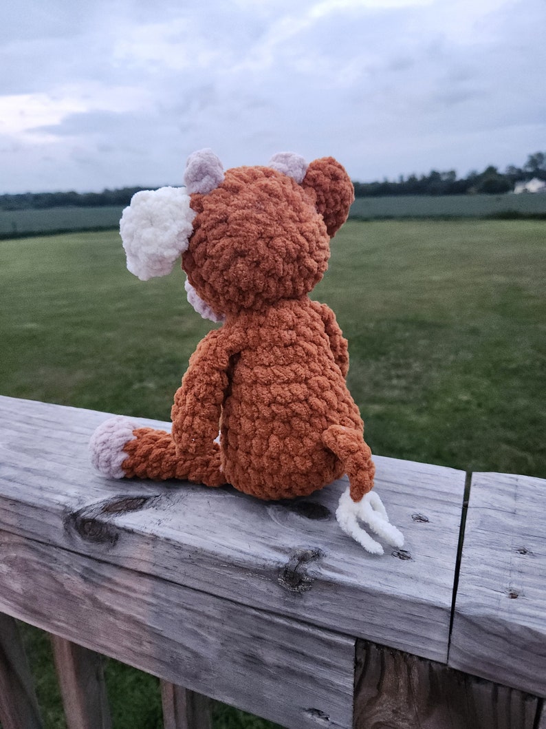 May include: A brown crocheted stuffed animal with white ears and a white tail sitting on a wooden railing. The animal is looking out at a field in the distance.