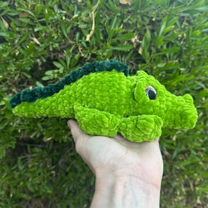 May include: A handmade crochet plush toy shaped like a small alligator. The body is made of bright lime green yarn with a textured, fuzzy appearance. It features a dark forest green ridge along its back and a single large, round black eye.