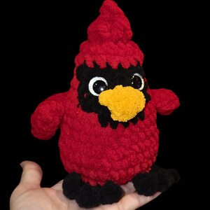 K&ouml;nnte beinhalten: Ein rotes, geh&auml;keltes Pl&uuml;schtier in Form eines Kardinals mit schwarzen Augen, einem schwarzen Schnabel und einem gelben Schnabel. Das Spielzeug sitzt auf einer Hand.