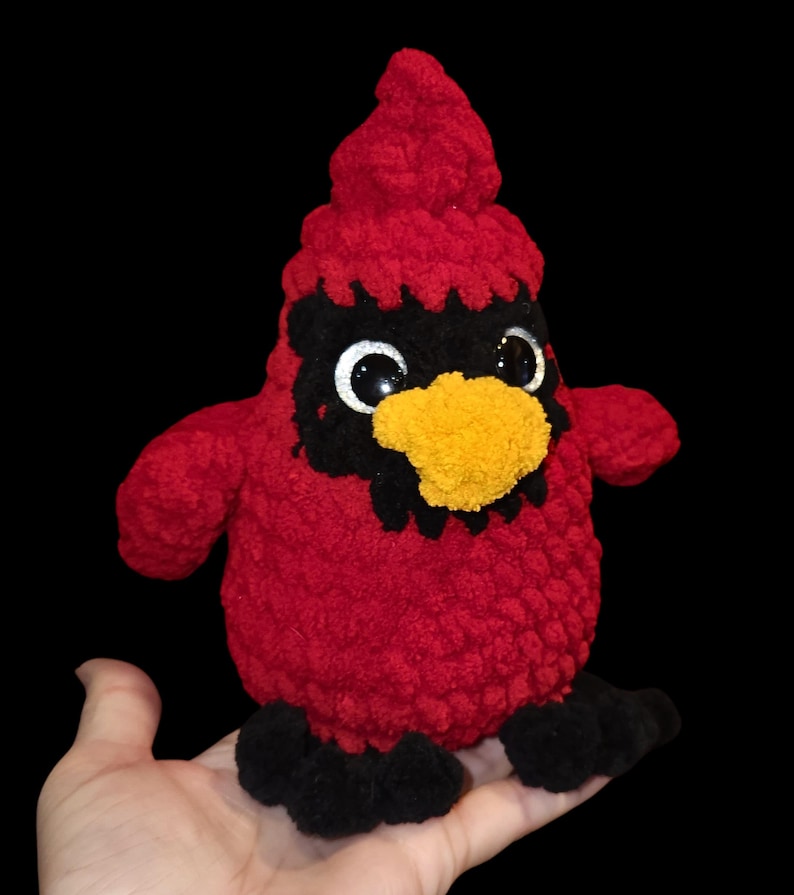 K&ouml;nnte beinhalten: Ein rotes, geh&auml;keltes Pl&uuml;schtier in Form eines Kardinals mit schwarzen Augen, einem schwarzen Schnabel und einem gelben Schnabel. Das Spielzeug sitzt auf einer Hand.