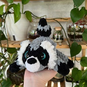 Op de afbeelding: Een pluche wasbeerspeeltje gemaakt van zacht grijs, zwart en wit garen. Het knuffeldier heeft een gestreepte staart, witte vacht rond het gezicht, een zwarte neus en grote, ronde, iriserend blauwe ogen. De textuur is pluizig en dik.