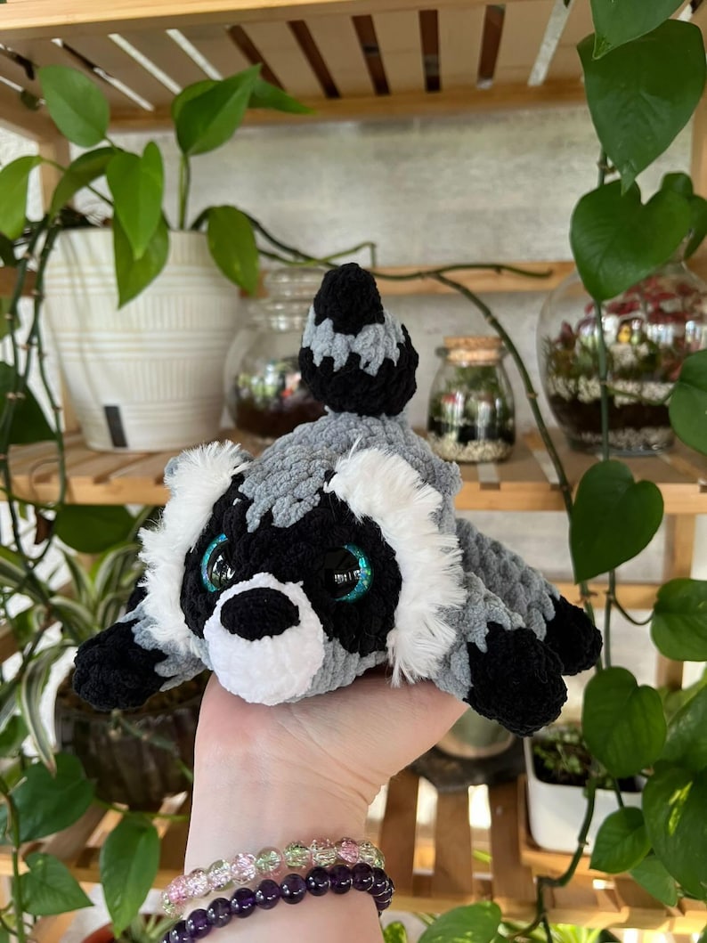 Op de afbeelding: Een pluche wasbeerspeeltje gemaakt van zacht grijs, zwart en wit garen. Het knuffeldier heeft een gestreepte staart, witte vacht rond het gezicht, een zwarte neus en grote, ronde, iriserend blauwe ogen. De textuur is pluizig en dik.