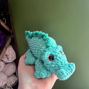 May include: A small, handmade crochet alligator toy crafted from soft, light turquoise yarn. The plush figure features a textured, bumpy spine, a rounded snout, and large, glossy black safety eyes with iridescent, glittery pink accents.