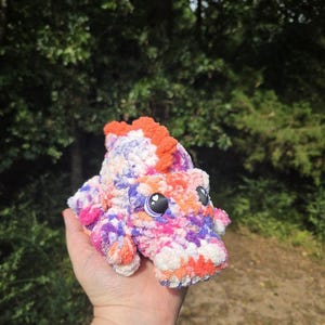 May include: A small, plush, crocheted dinosaur toy featuring a variegated yarn pattern in shades of orange, purple, pink, and white. The creature has a textured, fuzzy surface, a row of orange spikes along its back, and large, dark, glossy eyes.
