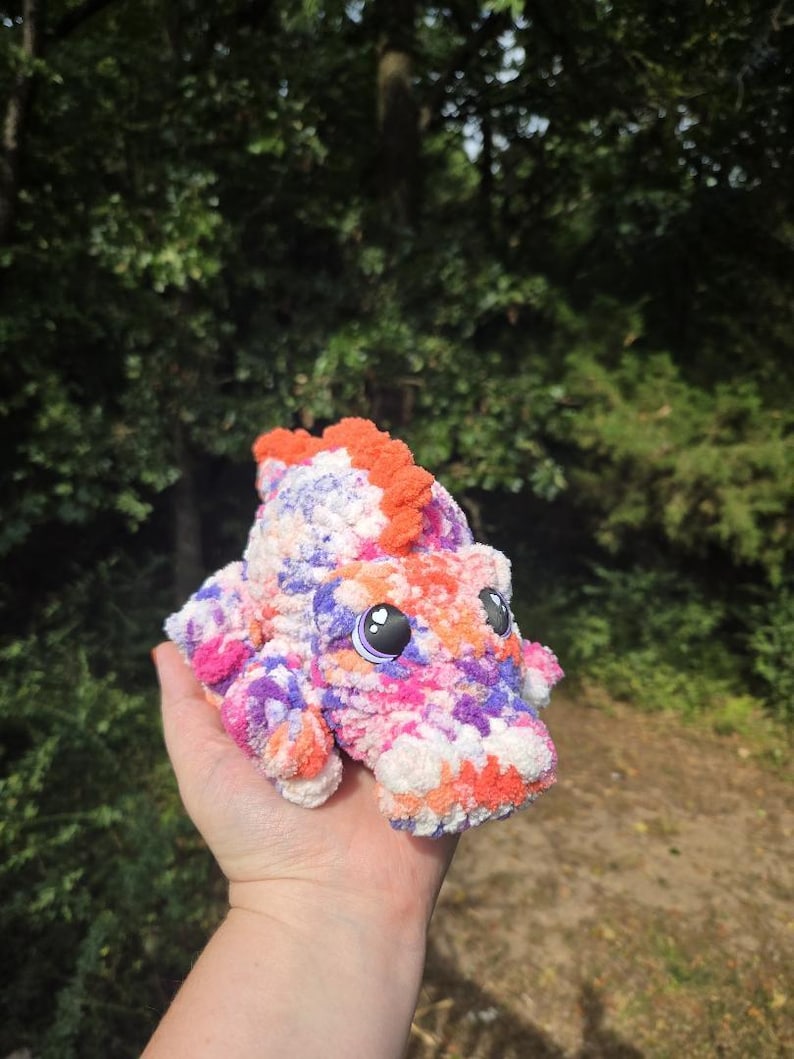 May include: A small, plush, crocheted dinosaur toy featuring a variegated yarn pattern in shades of orange, purple, pink, and white. The creature has a textured, fuzzy surface, a row of orange spikes along its back, and large, dark, glossy eyes.