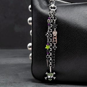 May include: A black textured handbag featuring a silver-toned metal chain charm. The charm includes a skull bead, a lime green star, purple beads, a silver spike, a zipper pull, a small cross, and a skull pendant embellished with green crystals.