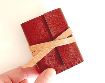 May include: A small, red leather journal with a tan leather strap tied around it.