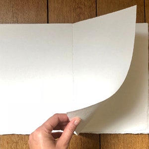 Artist turns the open page of a cotton rag khadi paper sketchbook with natural linen ribbon, Large size, which rests on a warm wood surface.