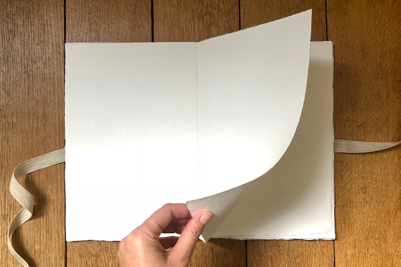 Artist turns the open page of a cotton rag khadi paper sketchbook with natural linen ribbon, Large size, which rests on a warm wood surface.