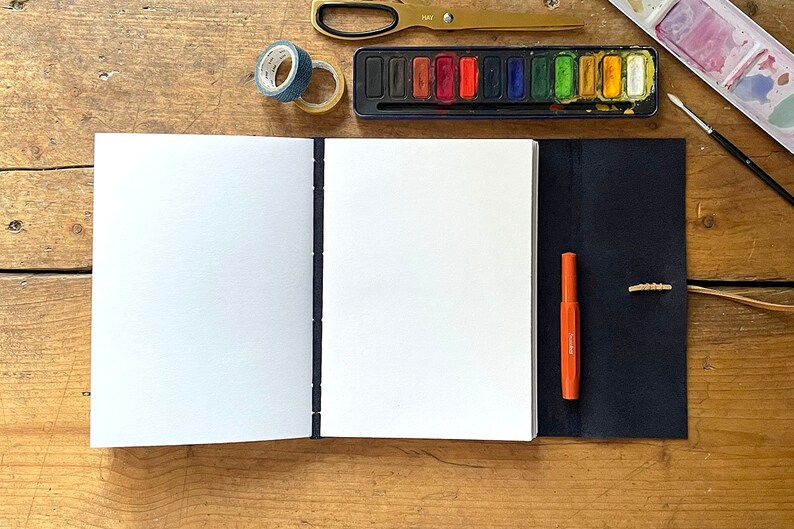 Inside pages of a Navy Blue sketchbook / art journal, showing its recycled cartridge pages. It is shown with watercolour paint palette, washi tape, brush, scissors and pen. It sits on a rustic wood surface.