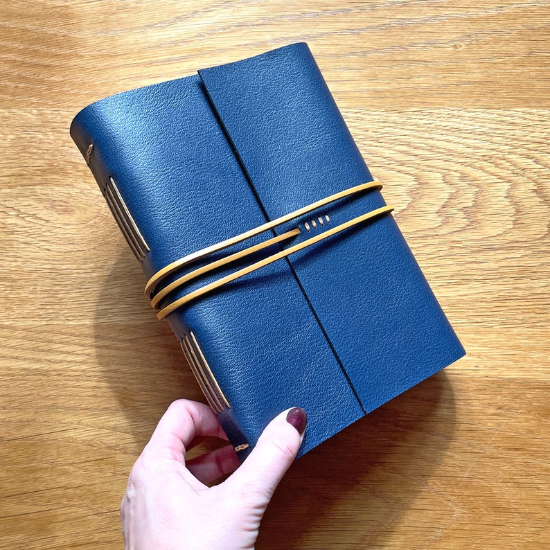 Bookbinder holds a Navy Blue Leather book bound in the Longstitch with Linkstitch exposed spine style in Tan linen thread, and with a Tan leather thong wrapping around the middle to keep it closed.