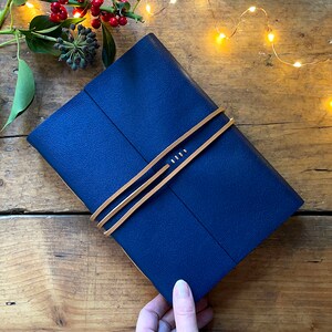 Leather Sketchbook in Navy Blue and Tan with thong and exposed spine lay flat binding. A Luxurious Christmas gift for artists. It is held at the corner and sits on a wood background, with holly and ivy berries and twinkly festive lights.