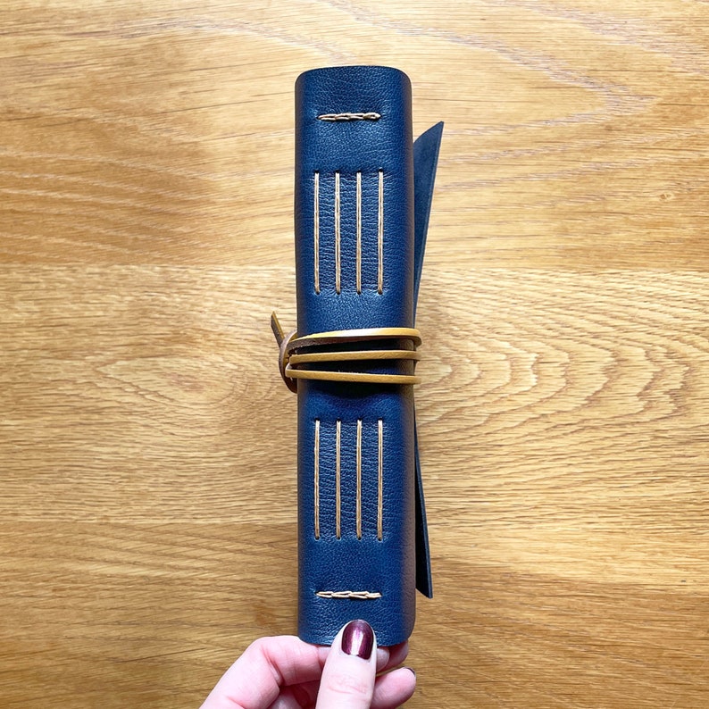 Medieval Leather Sketchbook bound in Navy Blue, with Longstitch with Linkstitch exposed spine turned to face the camera. A Tan leather thong wraps around its middle to keep it close. It sits on pale wood background.