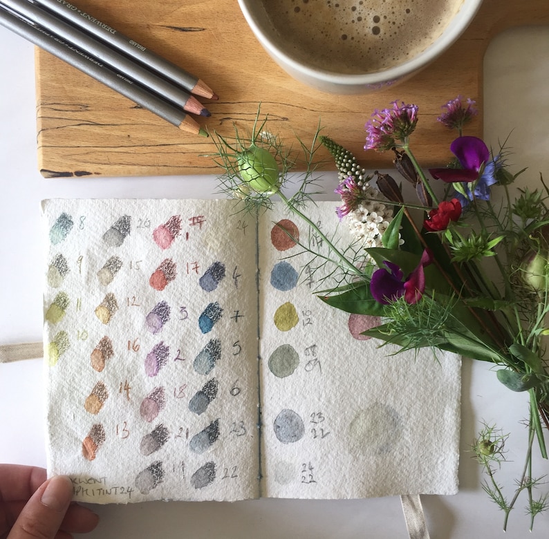 Cotton rag sketchbook with watercolour and pencil colour samples, lay flat khadi paper art journal gift for artists. Shown with a posy of garden flowers, coffee and artist pencils.