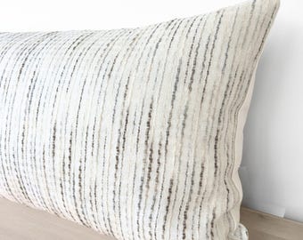May include: A rectangular throw pillow featuring a textured off-white fabric base with vertical stripes in varying shades of gray and brown. The woven material has a visible, slightly irregular grain, creating a subtle pattern across the surface.