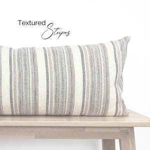 May include: A white and gray striped throw pillow with the text "Textured Stripes" on a white background. The pillow is sitting on a light wood bench.
