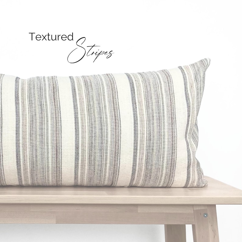 May include: A white and gray striped throw pillow with the text "Textured Stripes" on a white background. The pillow is sitting on a light wood bench.