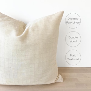 May include: A cream-colored, double-sided pillow with a plaid textured design. The pillow is made of raw linen and is dye-free.
