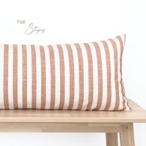 May include: A long, rectangular pillow with a brown and white striped pattern. The pillow is made of a soft, textured fabric and is sitting on a wooden bench. The text "Fall Stripes" is written in the top left corner of the image.