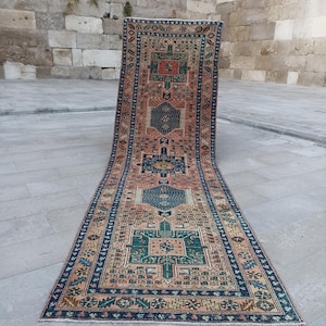 May include: A long, rectangular runner rug featuring a complex geometric pattern in shades of terracotta, deep blue, and forest green. The design includes multiple central medallions and a detailed border, with a short fringe at the ends.