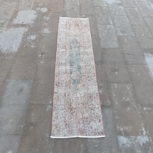 May include: A long, narrow rug with a floral pattern in shades of pink, white, and green. The rug has a fringed edge and is laid out on a gray tiled floor.
