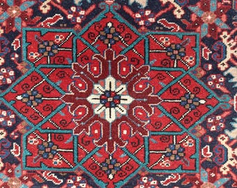 May include: Close-up of a detailed, patterned rug with a central geometric design. The rug features a rich color palette of red, navy blue, teal, and cream. Intricate floral and geometric motifs are woven throughout the rug's surface.