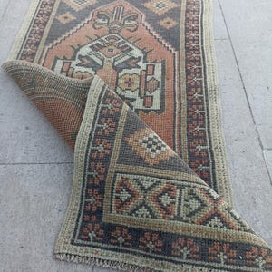 May include: A rectangular area rug featuring a central geometric medallion in shades of peach, charcoal gray, and cream. The rug has a wide border with repeating floral motifs and a fringed edge, with one corner folded over to reveal the woven backing.