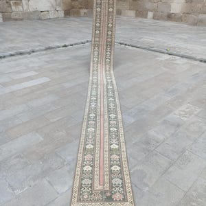 May include: A long, narrow runner rug with a detailed pattern. The rug features a central pink stripe flanked by patterned borders in shades of green, beige, and pink. The rug is displayed outdoors on a stone surface.