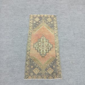 May include: Rectangular area rug with a central diamond pattern. The rug features a salmon-colored center, framed by a dark gray border with geometric designs. The rug's design includes beige and light green accents.
