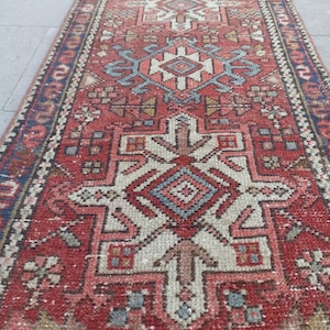 May include: A long, narrow runner rug featuring a deep red background with intricate geometric patterns in cream, light blue, and dark blue. The design includes repeating central diamond-shaped medallions and a detailed border along the edges.