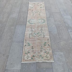 Könnte beinhalten: Ein rechteckiger Teppich im Vintage-Stil mit verblasstem Blumenmuster in Beige, Grün und Creme. Der Teppich hat ein zentrales Design mit sich wiederholenden Blumenmotiven und ein verwittertes Aussehen, geeignet für einen Flur oder Eingangsbereich.