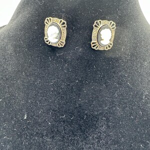 May include: A pair of gold-tone earrings with black and white cameo insets. The cameos are oval shaped and feature a profile of a person. The earrings have a detailed design with black accents.
