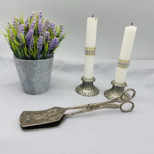 May include: A still life composition with a potted lavender plant, two white candles with rhinestone bands, and a silver serving utensil. The candles are in ornate silver holders. The serving utensil has a decorative floral pattern.