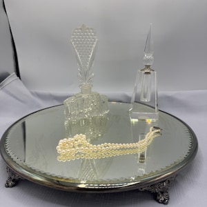May include: A vintage vanity set featuring a round mirrored tray with ornate silver-toned feet. The tray holds two clear glass perfume bottles and a strand of faux pearls. The bottles have decorative stoppers.