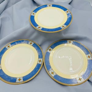 May include: Three white ceramic plates with a blue and yellow border. The blue border features a pattern of white stripes and small red and green floral accents. The plates have a gold trim.