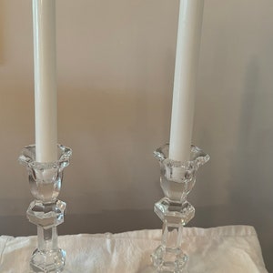 May include: Two clear glass candle holders with white candles. The candle holders have a geometric design and are made of crystal. The candle holders are on a white surface. The candle holder on the right has a label that says "Violetta Made in Poland"