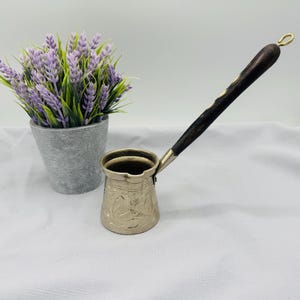 May include: A silver Turkish coffee pot with an ornate design and a dark wood handle. The pot has a small spout and a rounded base.
