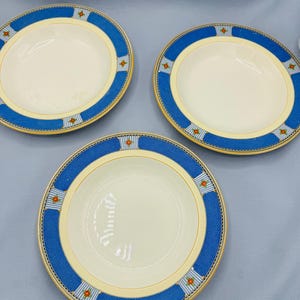 May include: Three cream-coloured ceramic bowls with a blue border. The border features a repeating pattern of red and white accents. The bowls have a wide rim and a shallow depth, suitable for serving soups or salads.